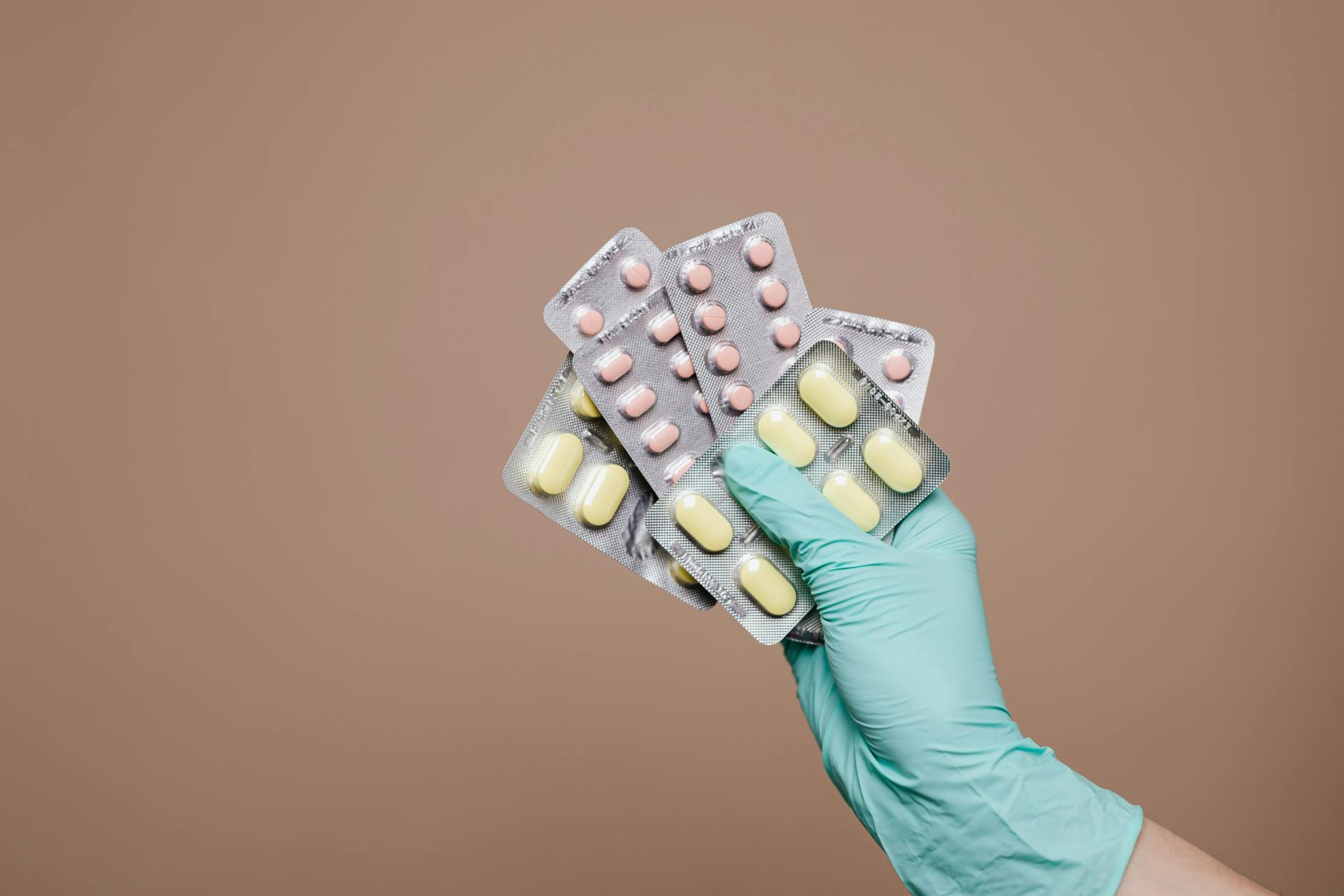 A gloved hand holding several blister packs of tablets against a plain brown background