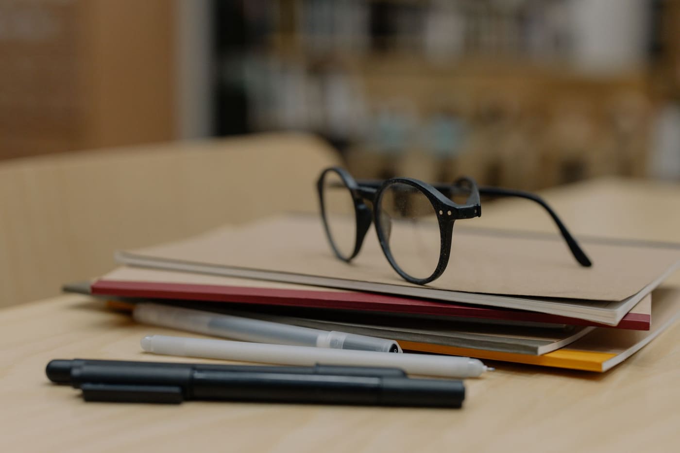 A pair of black-framed glasses resting on a stack of notebooks beside pens on a wooden desk