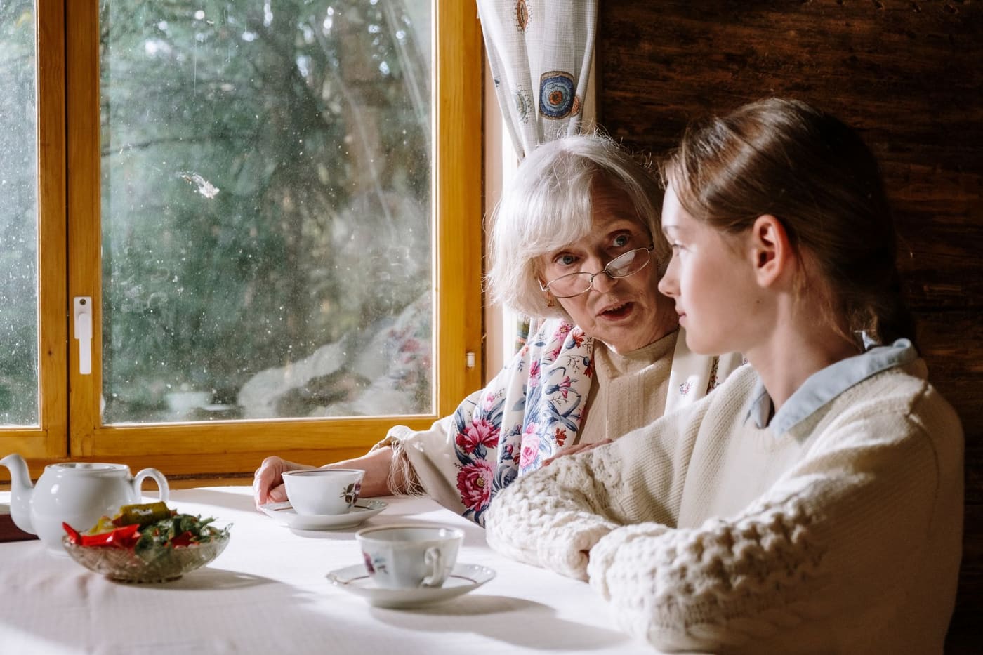 A grandmother and granddaughter sitting together at a window table with cups of tea