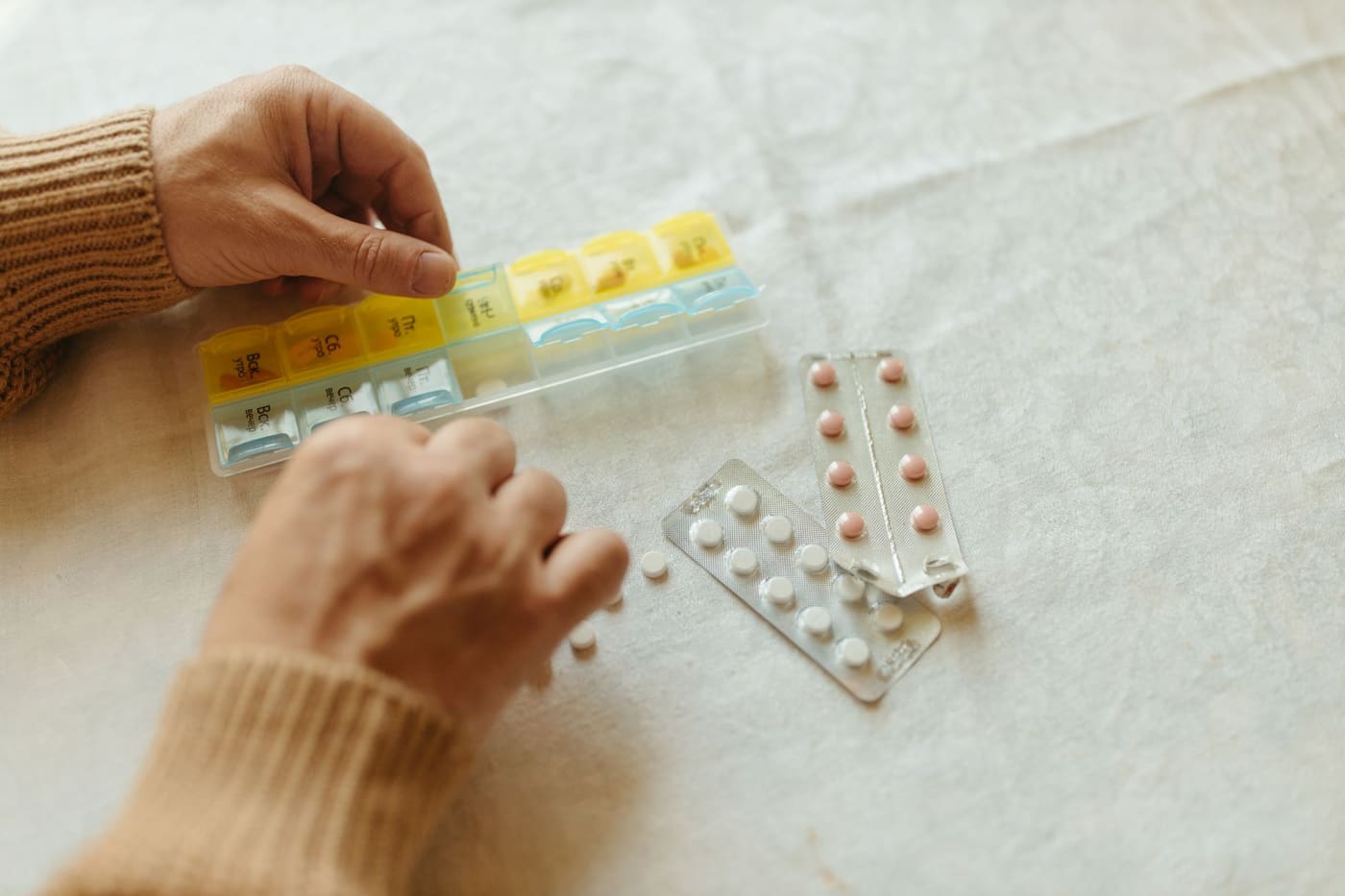 Two hands filling a yellow and blue weekly pill organizer beside strips of tablets