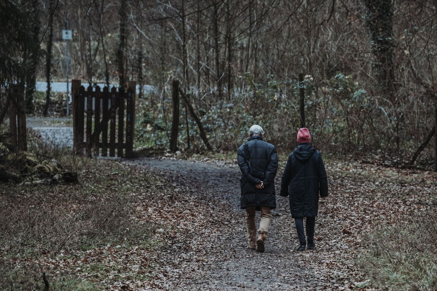 Two older adults in dark coats walking together along a leaf-covered path through a wood