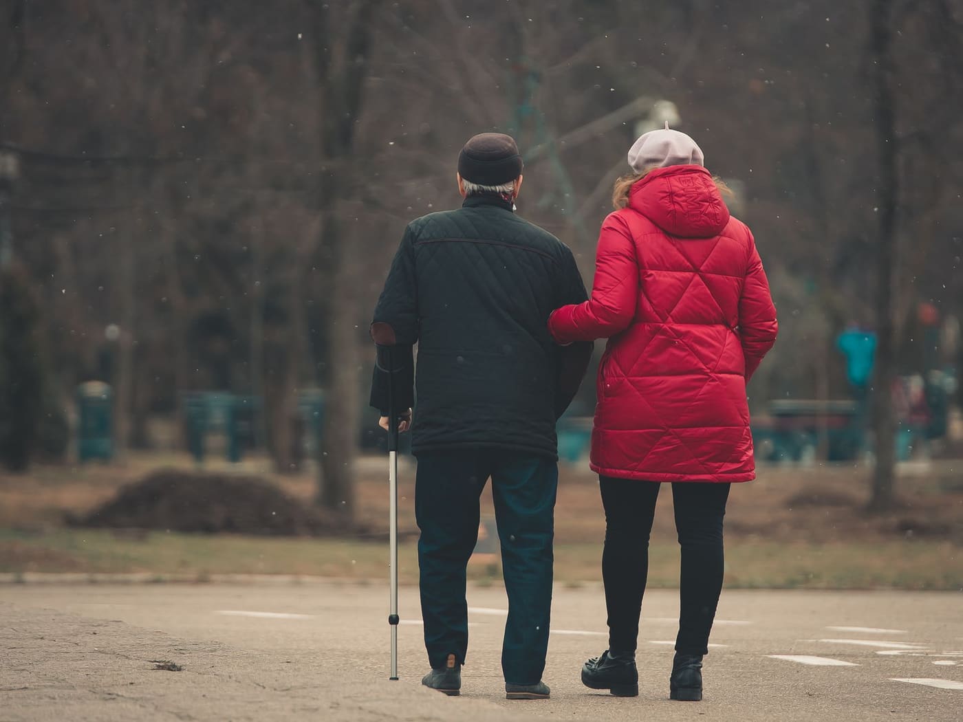 An older man with a walking stick and a woman in a red coat walking arm in arm along a park path