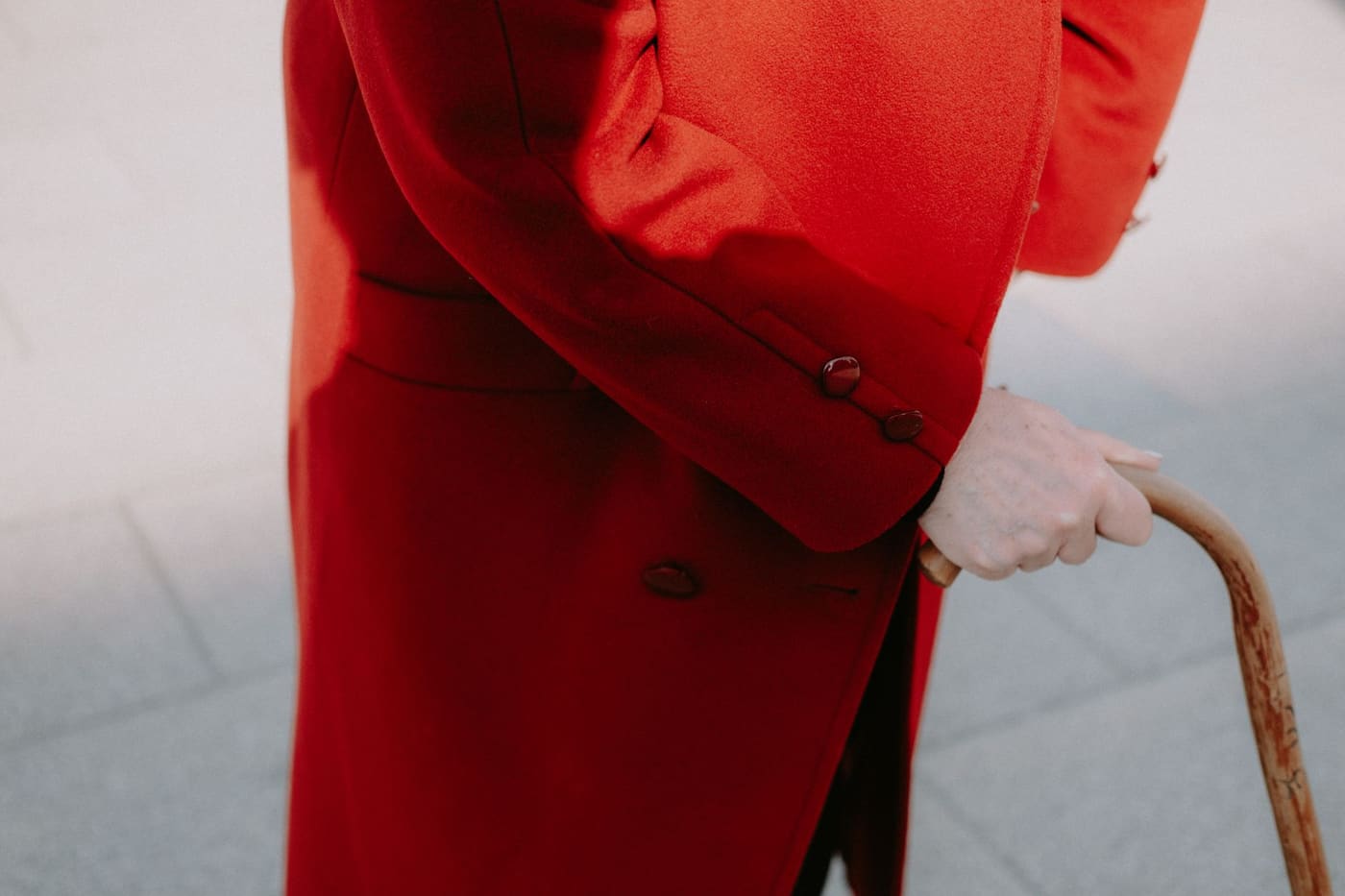 An older person in a long red coat holding the curved handle of a wooden cane
