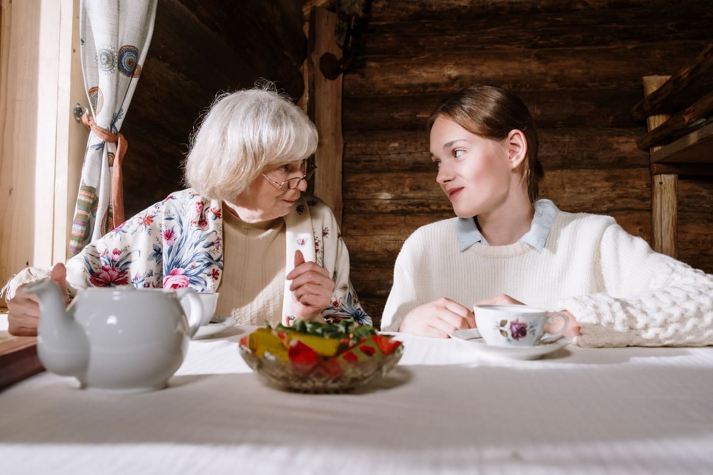 An older woman and a younger woman talking across a table set with a teapot and fruit bowl