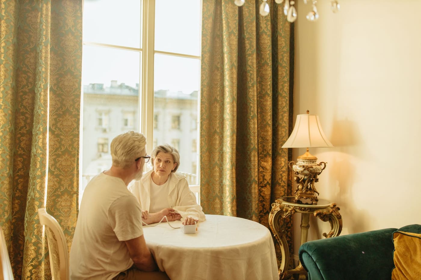 An older couple sit at a round table by a tall window while one takes the other's blood pressure with a home monitor