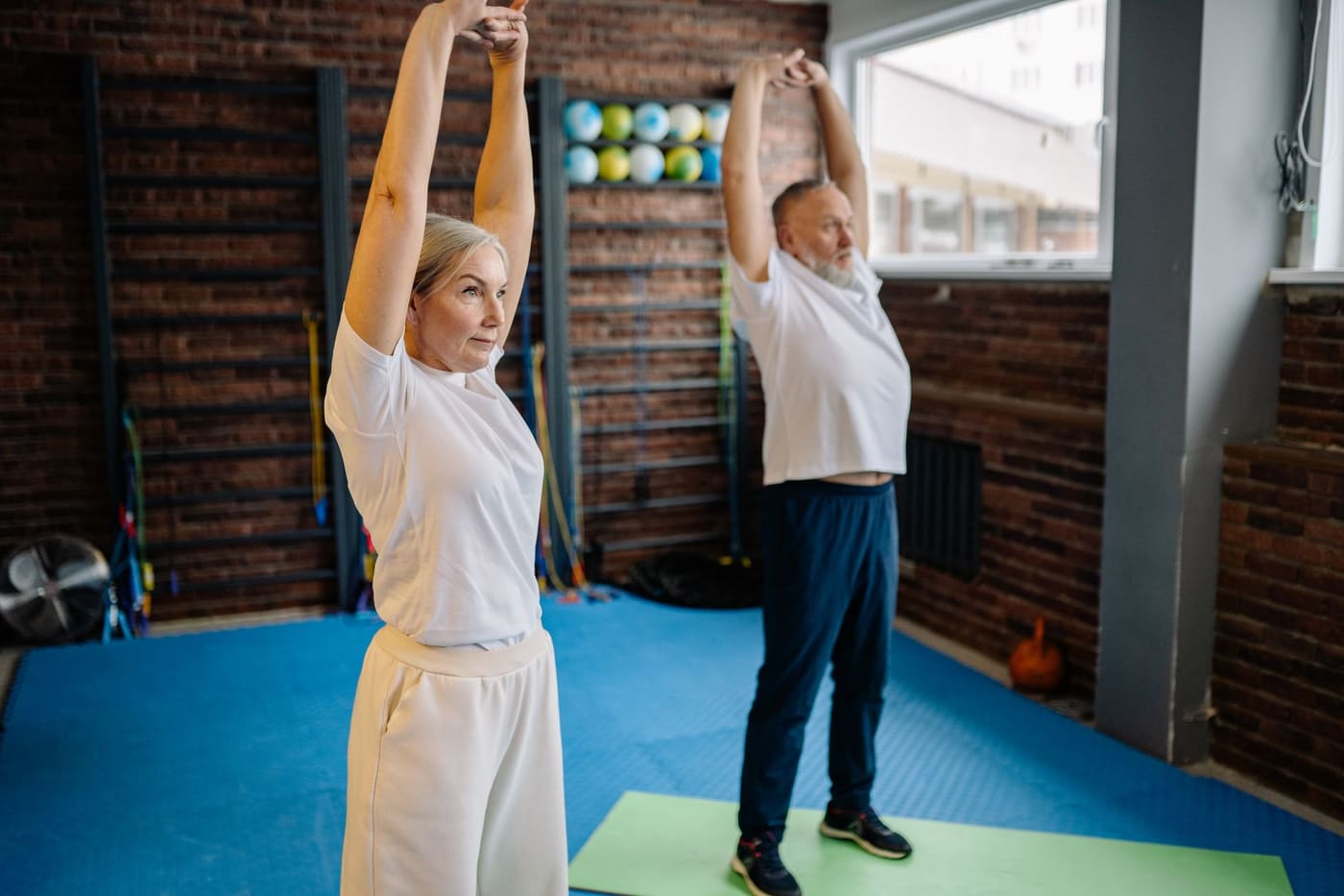Two older adults stretching with arms raised overhead on exercise mats in a bright room
